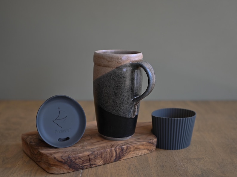 May include: A ceramic travel mug with a handle, a lid, and a silicone sleeve. The mug is two-toned, with a black base and a gray and tan upper section. The lid is dark gray and has a logo. The sleeve is dark gray.