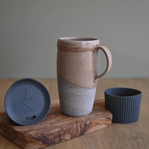 May include: A ceramic travel mug with a handle, a lid, and a silicone sleeve. The mug is two-toned, with a tan upper half and a gray lower half. The lid is gray with a logo. The sleeve is dark gray.
