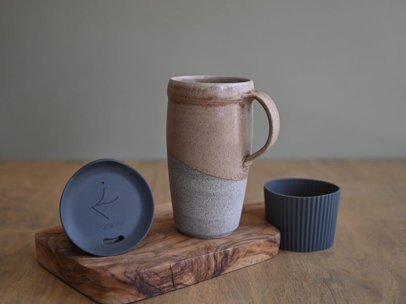 May include: A ceramic travel mug with a handle, a lid, and a silicone sleeve. The mug is two-toned, with a tan upper half and a gray lower half. The lid is gray with a logo. The sleeve is dark gray.