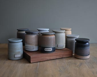 May include: A collection of nine cylindrical ceramic jars with matching lids, featuring various earth-toned glazes including gray, tan, brown, and black. Several jars have rectangular labels with text such as LENT, ELDERLY, and PORTER embossed on them.