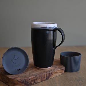 May include: A black ceramic travel mug with a white rim, a handle, and a matching lid. The lid and a ribbed sleeve are next to the mug on a wooden cutting board. The lid has the text "PortaVia".