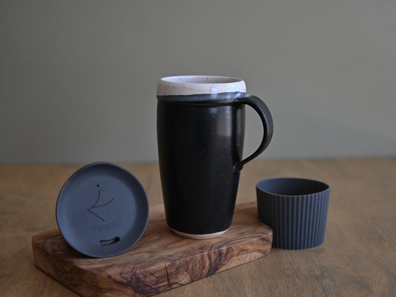 May include: A black ceramic travel mug with a white rim, a handle, and a matching lid. The lid and a ribbed sleeve are next to the mug on a wooden cutting board. The lid has the text "PortaVia".