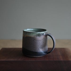 Könnte beinhalten: Eine handgefertigte Keramiktasse mit dunkler, strukturierter Oberfläche. Die Tasse hat einen Farbverlauf aus Schwarz, Braun und Grün am Rand. Sie hat einen abgerundeten Griff und steht auf einer Holzoberfläche.