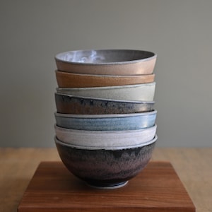 May include: A vertical stack of seven ceramic bowls featuring a variety of glazed finishes in shades of beige, tan, sage green, dark brown, light blue, white, and a deep charcoal black, each with a smooth, rounded rim and a consistent circular shape.