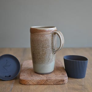May include: A ceramic travel mug with a handle, a lid, and a silicone sleeve. The mug is tan and brown with a speckled texture. The lid is dark blue, and the sleeve is ribbed and dark blue. The mug sits on a small wooden cutting board.