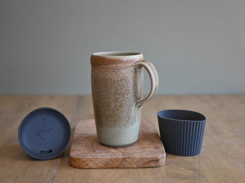 May include: A ceramic travel mug with a handle, a lid, and a silicone sleeve. The mug is tan and brown with a speckled texture. The lid is dark blue, and the sleeve is ribbed and dark blue. The mug sits on a small wooden cutting board.