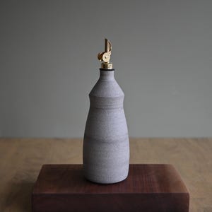 Peut inclure: Un distributeur d'huile en céramique gris clair mat avec un col effilé et une base large. La bouteille présente un bec verseur de couleur or métallique avec un mécanisme de couvercle à charnière sur le dessus, offrant un design fonctionnel.