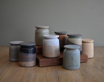 Può includere: Una collezione di otto barattoli in ceramica con coperchio, caratterizzati da vari smalti dai toni terrosi in sfumature di grigio, marrone, beige e blu. I barattoli sono disposti in gruppo, alcuni su un blocco di legno rettangolare, mostrando texture.