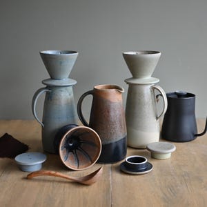 May include: A collection of ceramic coffee brewing equipment including two tall pitchers with matching cone-shaped drippers, a central brown and black pitcher, a black gooseneck kettle, two small ceramic lids, a wooden spoon, and a pile of coffee grounds.