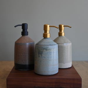 Op de afbeelding: Drie keramische zeepdispensers met een rustiek ontwerp. De dispensers zijn in tinten bruin, blauw en beige, met zwarte en gouden pompknoppen. Ze zijn gerangschikt op een donkerhouten dienblad.