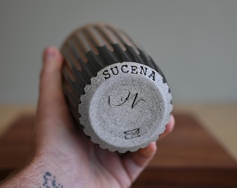 May include: A handmade ceramic tumbler with a textured, ribbed design. The top half is a dark gray, transitioning to a lighter tan. The bottom has the word "SUCENA" and artist's marks. The tumbler is held in a hand.