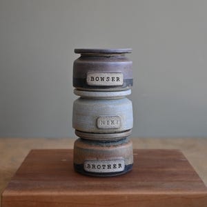 Op de afbeelding: Een stapel van drie cilindrische keramische potten met deksels. De bovenste pot is donkerbruin, de middelste is lichtgrijs en de onderste is beige. Elke pot heeft een rechthoekig label met de gestempelde tekst BOWSER, NIKI en BROTHER.