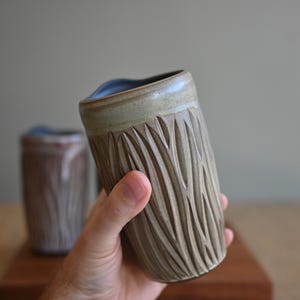 May include: A cylindrical ceramic tumbler with a light beige exterior featuring a carved vertical leaf-like pattern and a smooth, light blue interior glaze. The vessel has a slightly flared rim and a textured surface created by deep, rhythmic carvings.