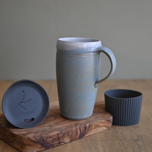 May include: A ceramic travel mug with a handle, a lid, and a ribbed sleeve. The mug is light blue with a white rim. The lid is dark gray and has the word "Fortiva" printed on it. The mug is on a wooden cutting board.