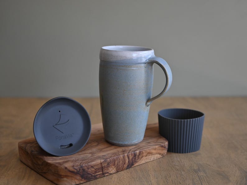 May include: A ceramic travel mug with a handle, a lid, and a ribbed sleeve. The mug is light blue with a white rim. The lid is dark gray and has the word "Fortiva" printed on it. The mug is on a wooden cutting board.