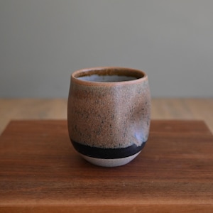 May include: A ceramic cup with a rounded, barrel-like shape. The exterior features a speckled brown and tan glaze on the upper portion, transitioning to a solid black band near the base, which is finished in a light, matte off-white color.
