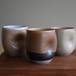 May include: Three ceramic cups with a rounded, handleless shape and a distinct thumb indentation on the side. The central cup features a dark brown base transitioning into a speckled tan glaze, while the other two cups display muted beige and brown tones.