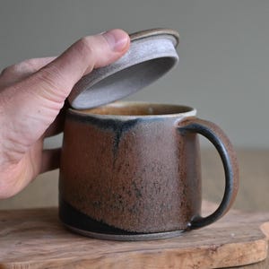 Könnte beinhalten: Handgefertigte Keramik-Tasse mit Deckel. Die Tasse ist braun mit schwarzen und blauen Akzenten und einem gebogenen Henkel. Der Deckel ist hellbraun. Die Tasse steht auf einem Holzbrett.