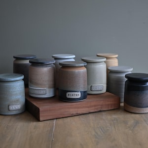 May include: A collection of ten cylindrical ceramic canisters with lids, featuring various earth-toned glazes including grey, brown, and black. Each canister has a small rectangular label with embossed text such as Porter and Lent.