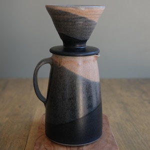 May include: A black and beige ceramic coffee pot with a matching ceramic pour-over filter. The pot has a handle and a spout. The filter sits on top of the pot.
