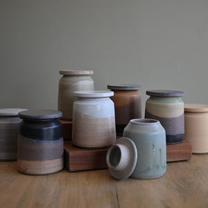May include: A collection of eight cylindrical ceramic jars with matching lids, featuring various earth-toned glazes including matte black, cream, sage green, and terracotta. The jars are arranged in a cluster, with one lid resting beside a jar.