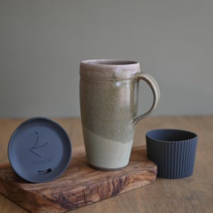 May include: A ceramic travel mug with a handle, in shades of beige and green, with a matching lid and a dark gray silicone sleeve. The mug sits on a wooden cutting board.