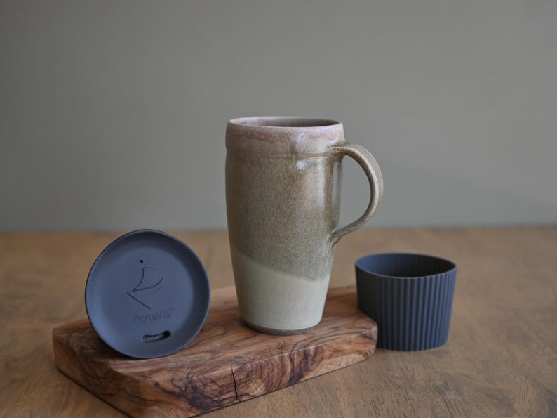 May include: A ceramic travel mug with a handle, in shades of beige and green, with a matching lid and a dark gray silicone sleeve. The mug sits on a wooden cutting board.