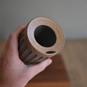 May include: A cylindrical ceramic tumbler with a dark grey, vertically ribbed exterior and a contrasting textured, speckled brown rim. The vessel features a circular opening at the top with a small, narrow horizontal slit near the edge of the rim.