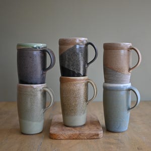 May include: A collection of six handcrafted ceramic mugs in various earth-tone colors. The mugs have a cylindrical shape with a handle and a slightly textured surface. Some mugs feature a two-tone design, and they are stacked on a wooden surface.