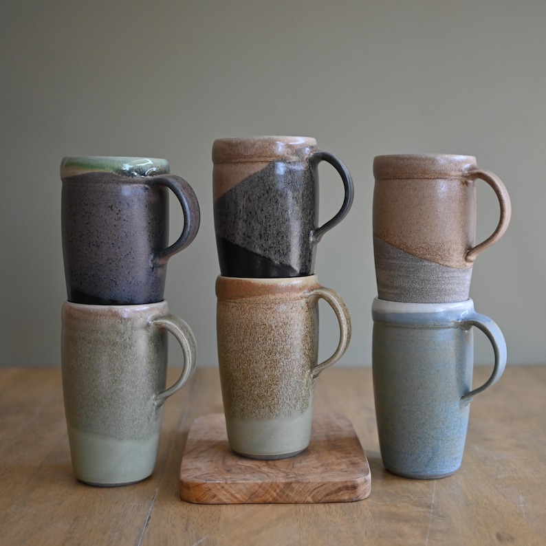 May include: A collection of six handcrafted ceramic mugs in various earth-tone colors. The mugs have a cylindrical shape with a handle and a slightly textured surface. Some mugs feature a two-tone design, and they are stacked on a wooden surface.