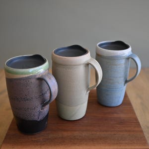 Peut inclure: Trois tasses de voyage en céramique avec couvercles et anses noirs. La tasse de gauche est violet foncé et noire avec un bord vert. La tasse centrale est beige clair avec une finition lisse. La tasse de droite est bleu clair texturé.