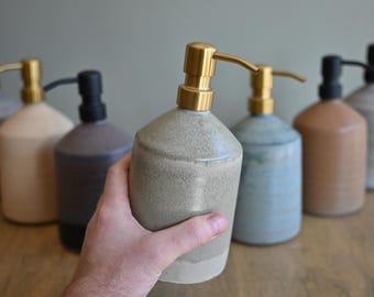 May include: A selection of ceramic soap dispensers in shades of grey, beige, blue, and brown. Each dispenser features a gold or black pump. One dispenser is held, highlighting the product's form and design.