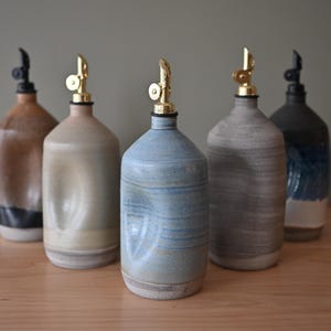 Può includere: Cinque dosatori per olio in ceramica con lati rientranti e beccucci metallici. Le bottiglie presentano smalti di vari colori tra cui marrone, beige, azzurro, grigio e una combinazione di blu scuro e bianco con finiture.