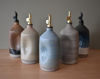 May include: Five ceramic oil dispensers with indented sides and metallic pour spouts. The bottles feature varied glaze colors including brown, beige, light blue, grey, and a dark blue and white combination, each with a distinct finish.