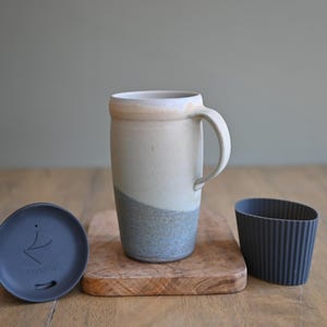 May include: A ceramic travel mug with a handle, a lid, and a sleeve. The mug is off-white with a blue-grey band at the bottom. The lid and sleeve are dark grey. The mug sits on a small wooden cutting board.