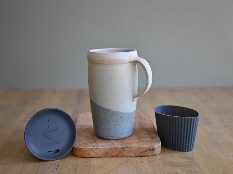 May include: A ceramic travel mug with a handle, a lid, and a sleeve. The mug is off-white with a blue-grey band at the bottom. The lid and sleeve are dark grey. The mug sits on a small wooden cutting board.