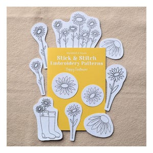 May include: A yellow rectangular card labeled Stick & Stitch Embroidery Patterns Happy Sunflowers, surrounded by various white fabric patches featuring black line art of sunflowers and rain boots with flowers, arranged on a tan surface.