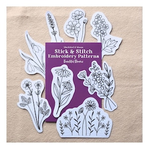 May include: A purple rectangular package labelled Stick & Stitch Embroidery Patterns Beautiful Blooms surrounded by eight white adhesive paper sheets featuring black line art illustrations of various wildflowers and botanical floral arrangements.