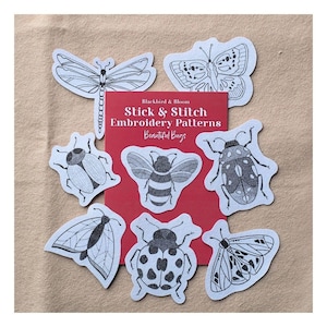 May include: A red rectangular card labelled Blackbird & Bloom Stick & Stitch Embroidery Patterns Beautiful Bugs, surrounded by eight white die-cut stickers featuring black line art illustrations of various insects including a dragonfly, bee, and beetles.