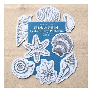May include: A blue rectangular card labelled blackbird & bloom Stick & Stitch Embroidery Patterns Seashells, surrounded by various white paper cutouts featuring black ink illustrations of a starfish, conch, and assorted seashells.