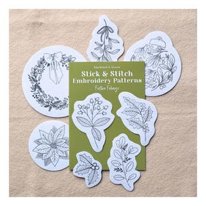 May include: A set of white stick and stitch embroidery patterns featuring black line art of festive foliage, including a wreath, mistletoe, ornament, poinsettia, and holly leaves, arranged around a green card labelled Stick & Stitch Embroidery Patterns.