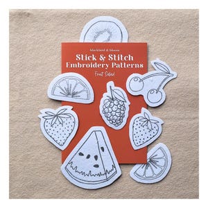 May include: A rectangular orange card labelled Stick & Stitch Embroidery Patterns Fruit Salad, surrounded by white fabric patches featuring black line art of a kiwi, orange slice, cherry, raspberry, strawberry, and watermelon slice.