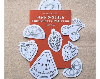 May include: A rectangular orange card labelled Stick & Stitch Embroidery Patterns Fruit Salad, surrounded by white fabric patches featuring black line art of a kiwi, orange slice, cherry, raspberry, strawberry, and watermelon slice.