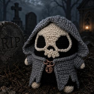 May include: A crocheted plush figure featuring a skull-shaped head with large black eye sockets, wearing a textured gray hooded cloak. The cloak is fastened at the neck with a metallic copper-colored chain and a circular lock pendant.