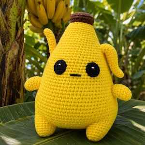 Puede incluir: Un peluche de ganchillo de color amarillo brillante con forma de plátano y un tallo marrón en la parte superior. La figura presenta dos grandes ojos circulares negros con pequeños reflejos blancos y una sencilla línea negra horizontal como boca.