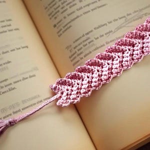 May include: A handmade, pink crocheted bookmark with a leaf-like design. The bookmark is lying on the open pages of a book. The bookmark has a tassel at the end. The color of the bookmark is a dusty rose color.