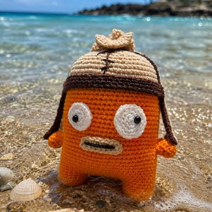 May include: A bright orange crocheted creature with two large white circular eyes, black pupils, and a tan mouth. The figure wears a tan and dark brown crocheted aviator-style hat with a small top knot and dark brown stitched detailing.