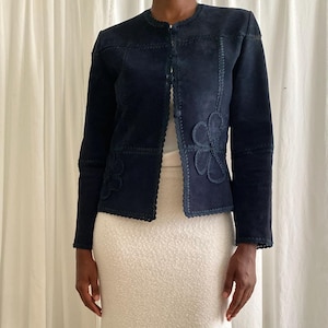 May include: A navy blue suede jacket with floral appliqués and scalloped edges. The jacket has a round neckline and a button closure. The model is wearing a cream-coloured textured skirt.