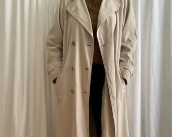 Könnte beinhalten: Beigefarbener Trenchcoat mit doppelreihigem Design. Der Mantel hat einen Reverskragen, Knopfverschlüsse und eine lange Länge. Das Model trägt ein braunes Hemd, eine schwarze Hose und braune Stiefel. Der Hintergrund ist ein weißer Vorhang.