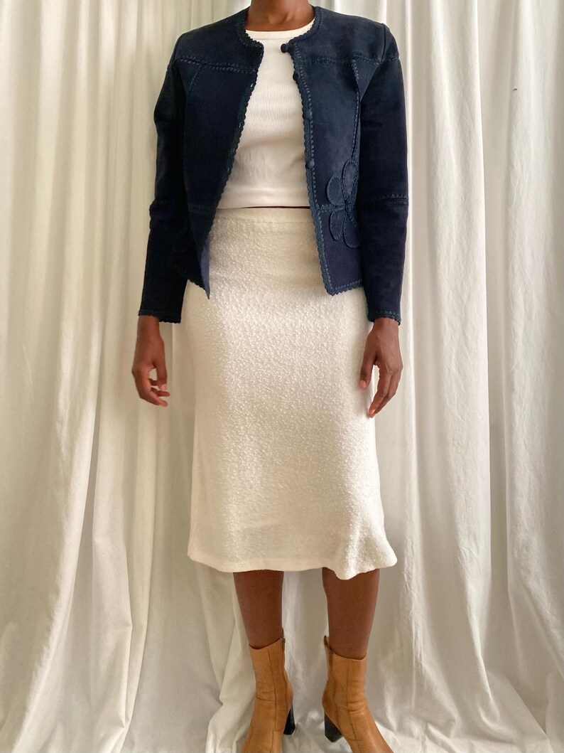 K&ouml;nnte beinhalten: Marineblaue Wildlederjacke mit floralen Applikationen und wei&szlig;em, strukturiertem Rock. Die Jacke hat einen runden Ausschnitt und lange &Auml;rmel. Der Rock ist knielang und das Model tr&auml;gt braune Stiefel.